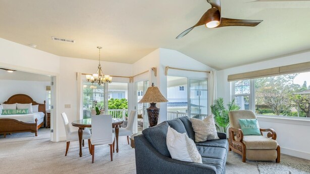 A spacious living area with a large gray sofa, a dining table with chairs, a ceiling fan, and a view of the outdoors. - Kauai Plantation At Princeville 1121 3 Bedroom Condo (Princeville)