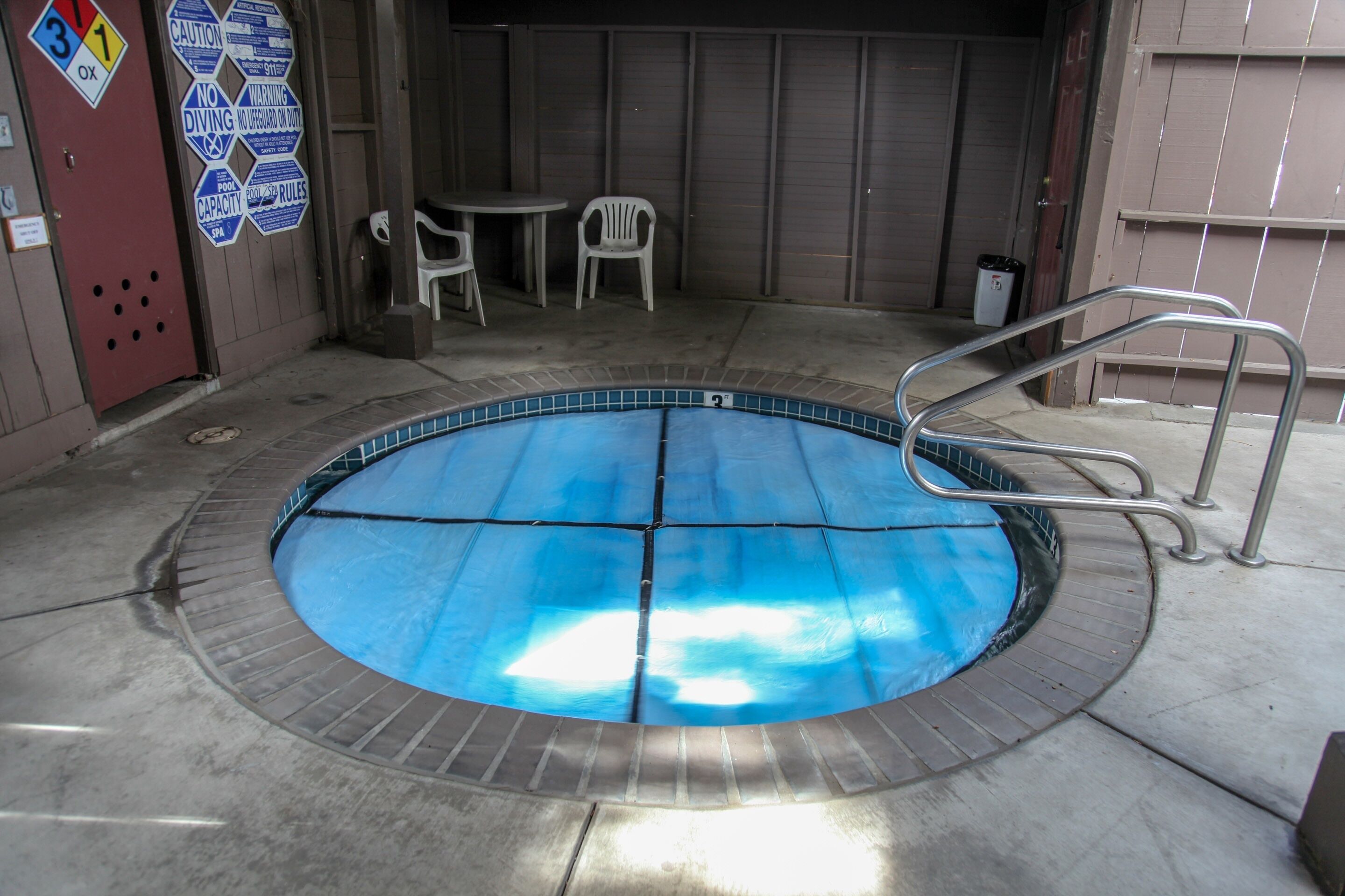 Piscina com escada metálica, rodeada por um deck de concreto e um espaço coberto com área de estar, cadeiras e mesa.