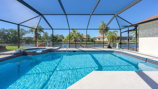 Ein Swimmingpool mit Glasveranda, umgeben von einer Terrasse mit Liegestühlen und Blick auf See und Bäume.