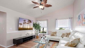 Ein Wohnzimmer mit weißem Sofa, einem Glastisch, einem Deckenventilator, einem an der Wand befestigten Fernseher und einem großen Fenster mit Jalousien.