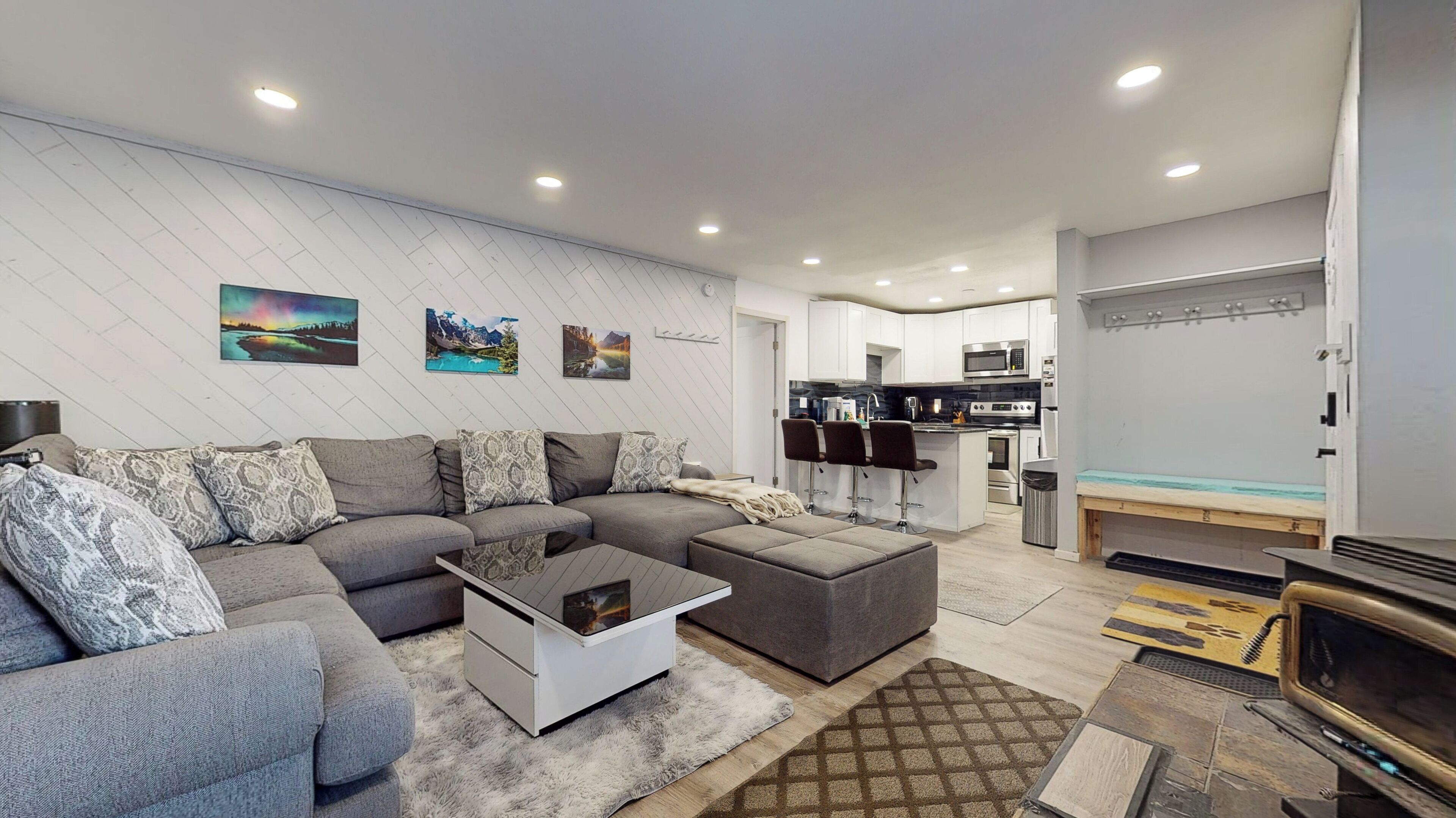 A modern living room with a grey sectional sofa, a white coffee table, and a TV. There is a kitchen area with white cabinets and a microwave. The room has a white wall with three landscape paintings.