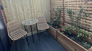 Una terraza de madera con dos sillas de mimbre y una mesita pequeña, rodeada por un cerco de madera y un macizo de plantas y flores.