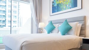 Een hotelkamer met een groot bed, twee blauwgroene kussens, een grijs hoofdeinde, een witte nachtkastje met een lamp, en een lijst met een abstract schilderij aan de muur.