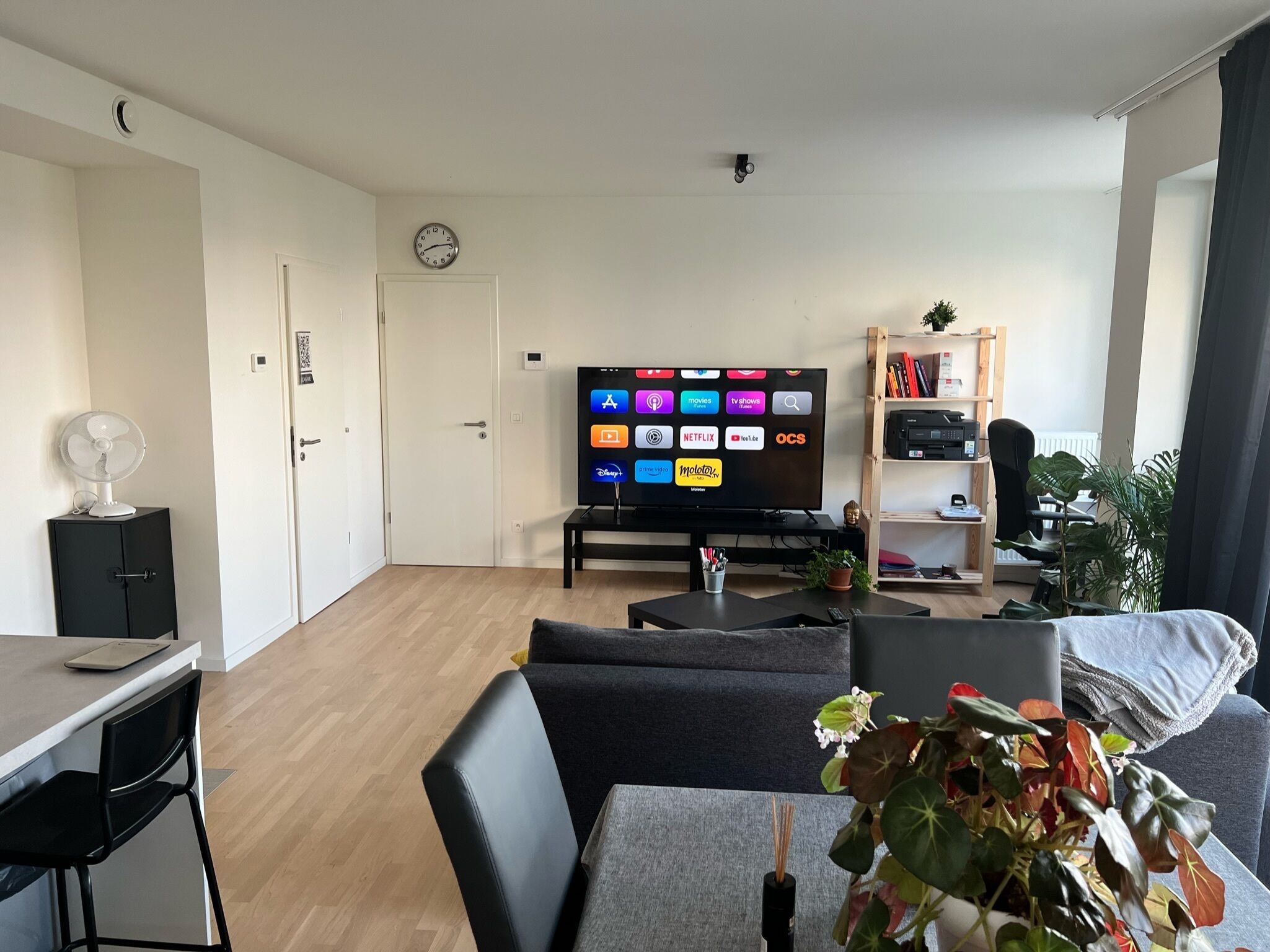 Ein modernes Wohnzimmer mit Flachbildfernseher, einem Essbereich mit Tisch und Stühlen, einer schwarzen Couch und einem kleinen Schreibtisch mit Ventilator.