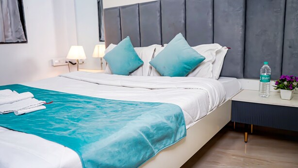 Un letto rifatto con copriletto color petrolio e lenzuola bianche, un comodino con una bottiglia d'acqua e un vaso di fiori, e una moderna testiera.