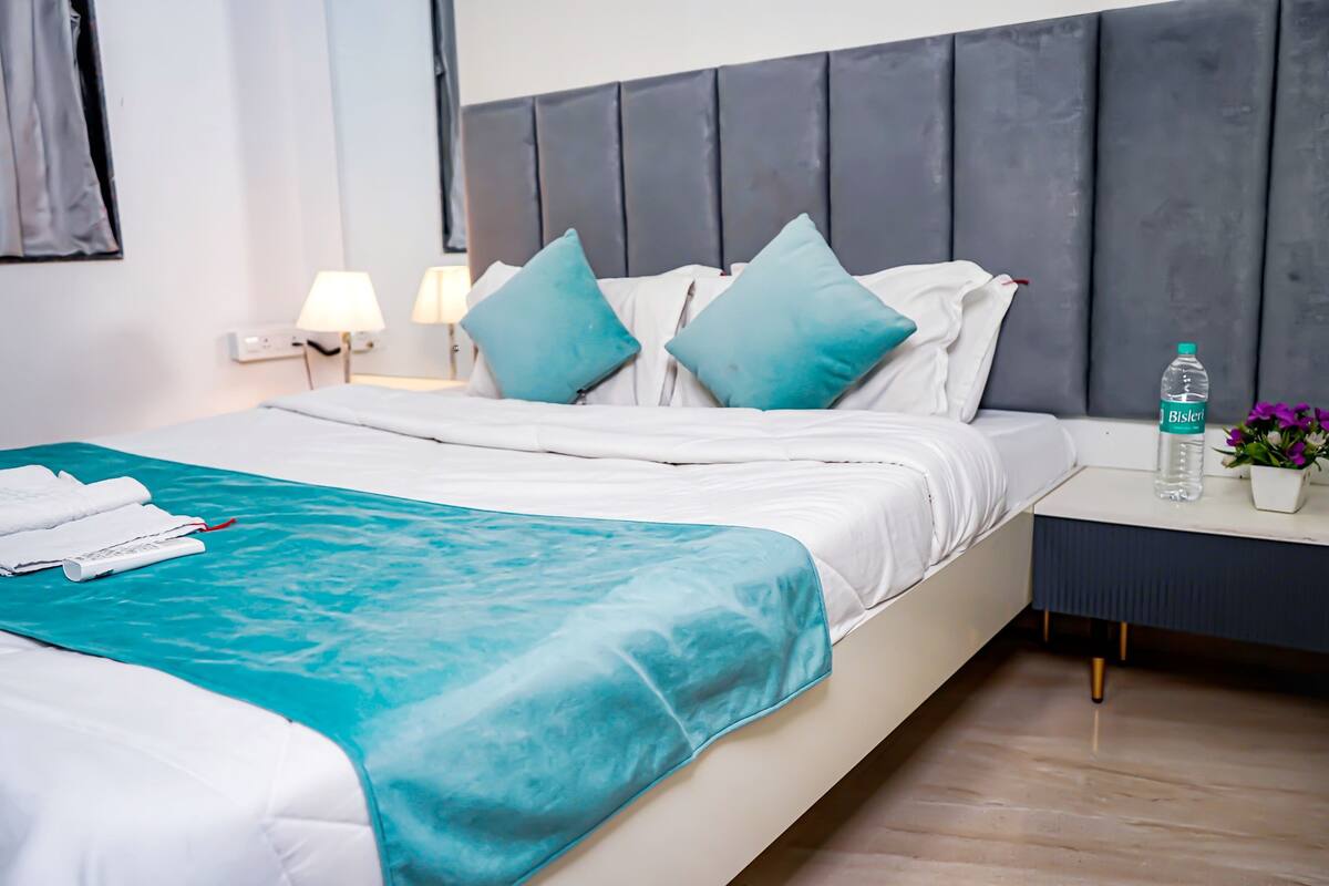 Cama bem arrumada com colcha azul-turquesa e lençóis brancos, mesa de cabeceira com uma garrafa de água e um vaso de flores, e cabeceira moderna.