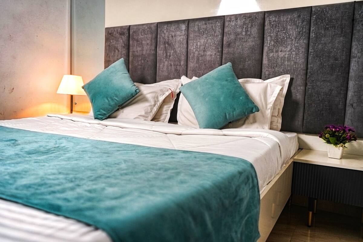 Cama bem arrumada com colcha e almofadas azul-turquesa, abajur e uma pequena planta em vaso sobre a mesa de cabeceira.