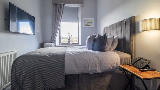 Une chambre à coucher avec un lit, une table de chevet avec un téléphone, une fenêtre avec des rideaux, un téléviseur fixé au mur et un radiateur.