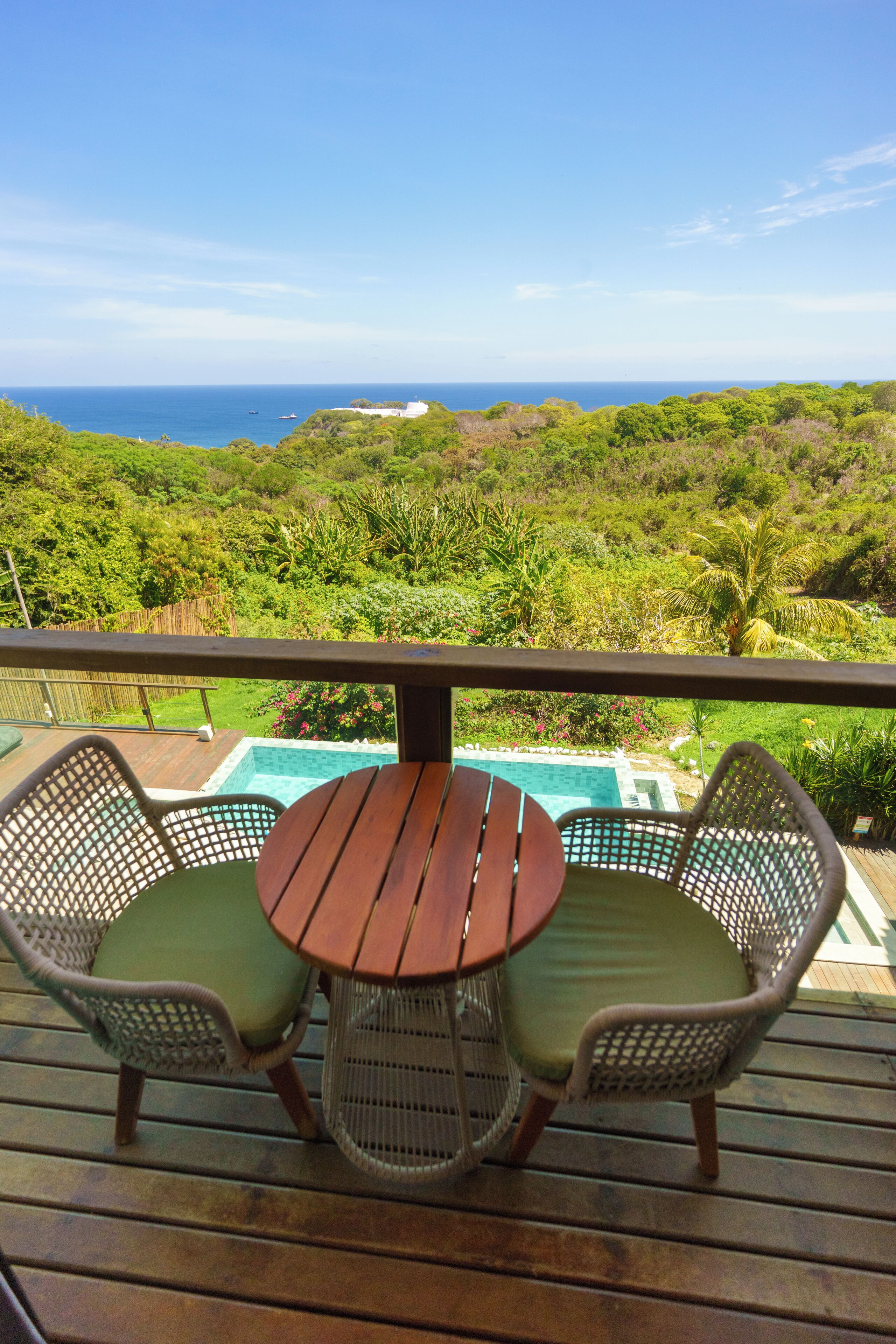 A wooden deck with a round table and two wicker chairs, overlooking a lush green landscape and a swimming pool.