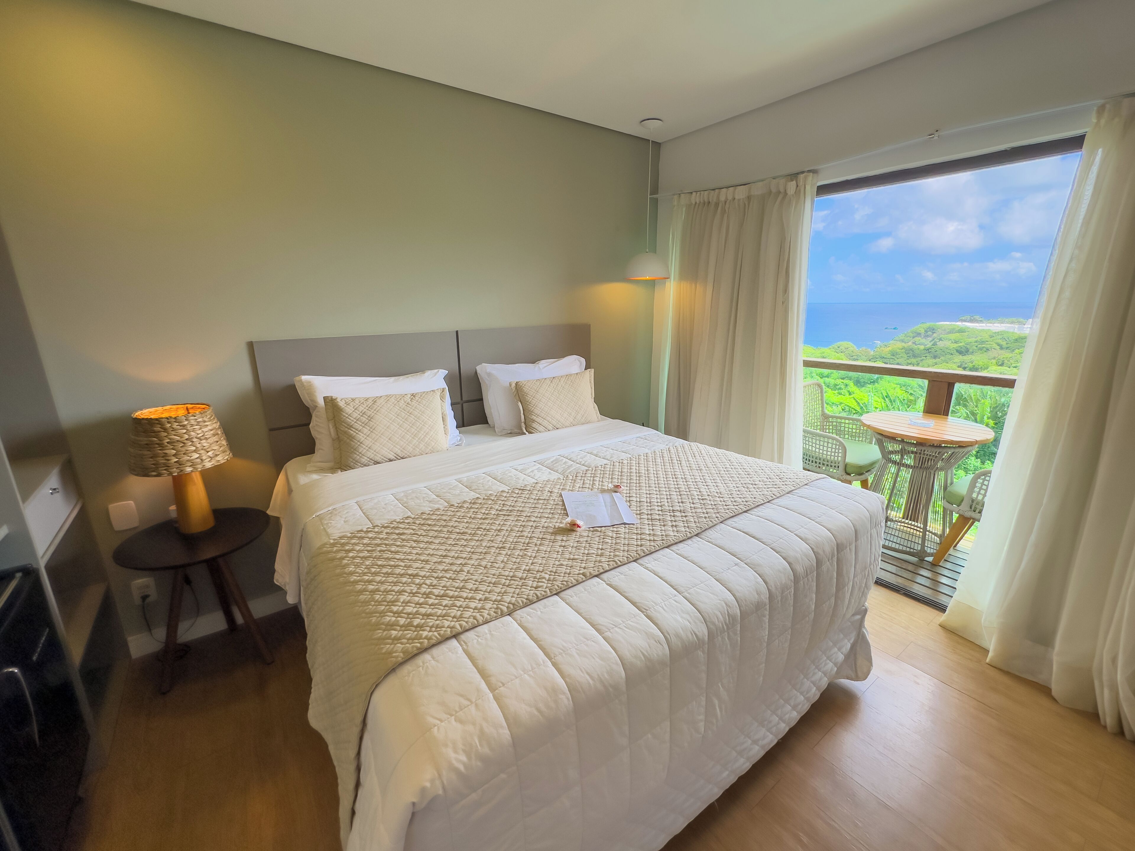 A hotel room with a large bed, a bedside table with a lamp, and a balcony with a view of the ocean.