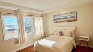 A bedroom with a large bed, a window with curtains, a nightstand, a painting on the wall, and a wooden floor. - Waterview Oasis by Buckroe Beach (Hampton)