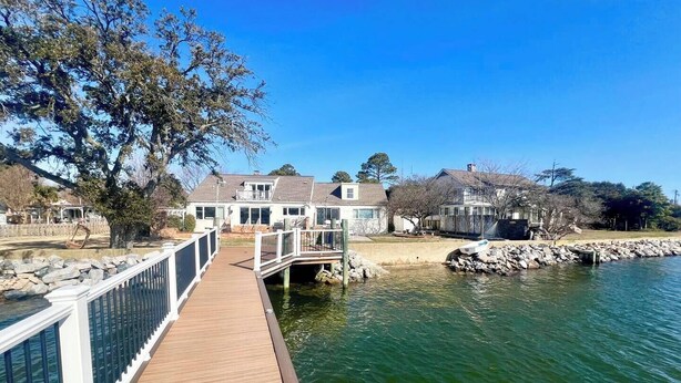 A wooden dock leading to a house by a calm body of water, with a stone wall and a tree in the foreground. - Waterview Oasis by Buckroe Beach (Hampton)