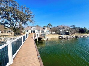 A wooden dock leading to a house by a calm body of water, with a stone wall and a tree in the foreground.