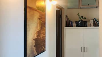Un pasillo con un cuadro grande de una escena de playa, un armario blanco lleno de libros y luces fijadas a la pared.