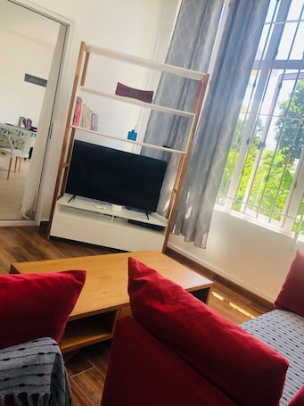 A living room with a TV on a stand, a wooden coffee table, red chairs, and a window with curtains.. Chez Moi