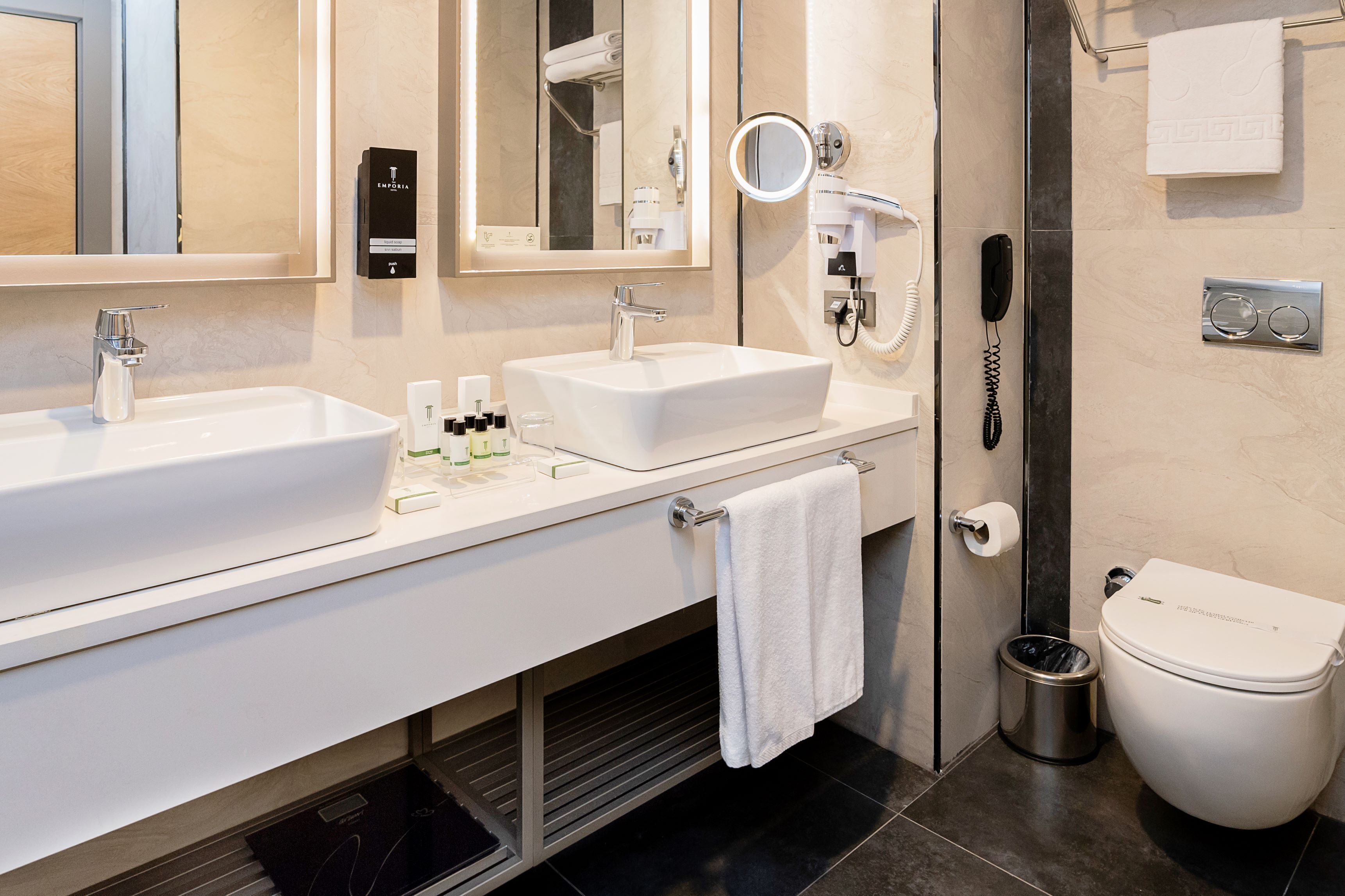 Une salle de bain moderne équipée de deux lavabos, d’un miroir, d’un sèche-cheveux, de toilettes et d’une poubelle.