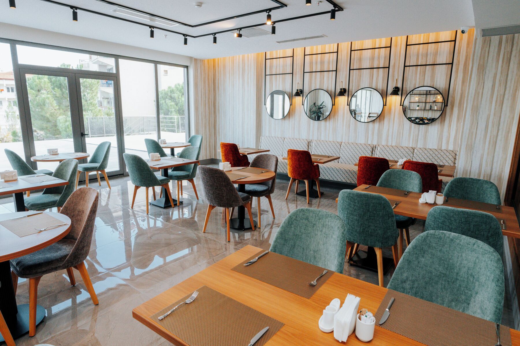 A modern restaurant with a mix of round and rectangular tables, green and red upholstered chairs, and a patterned wall with mirrors.