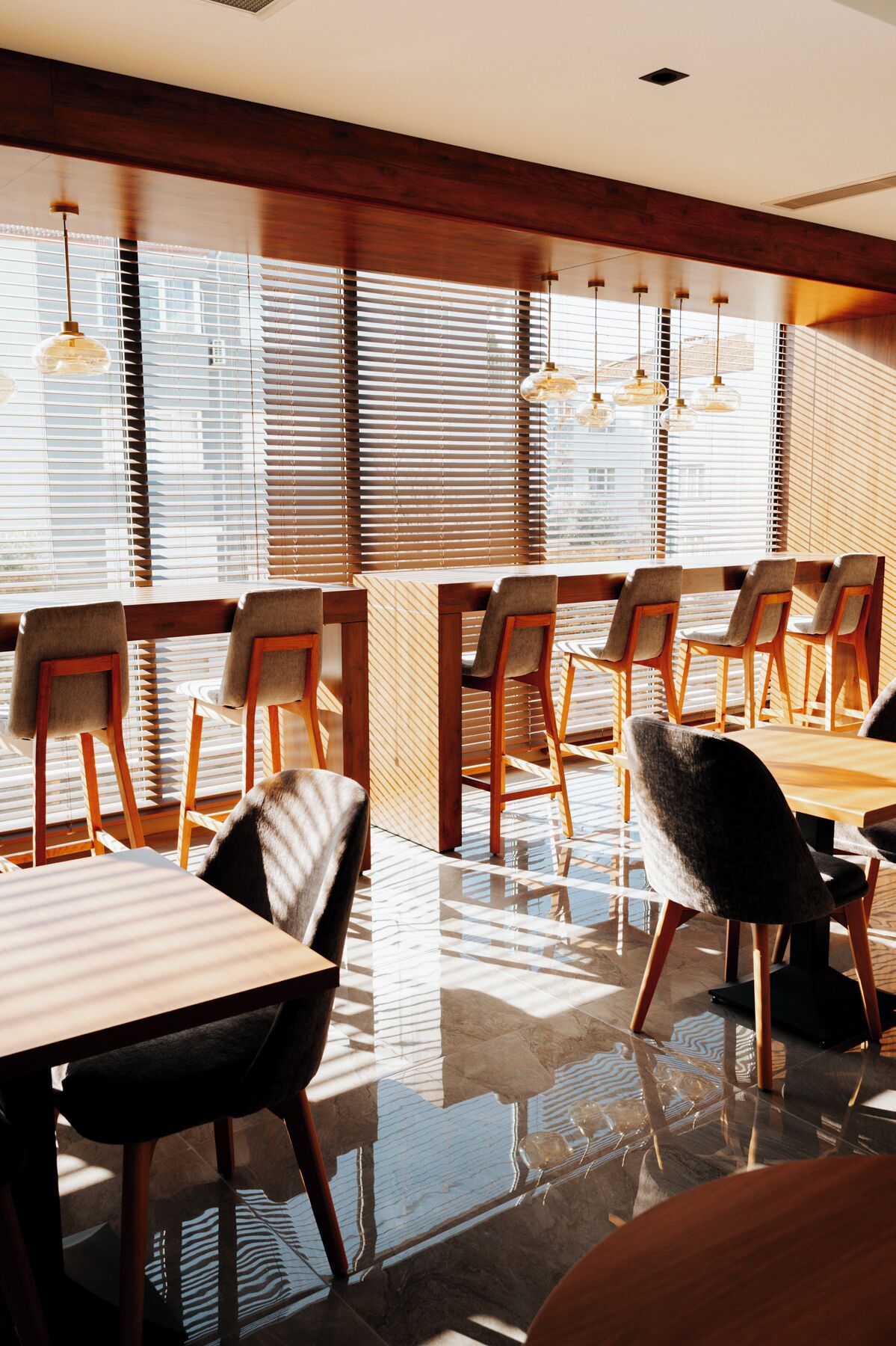 A modern dining area with wooden tables and chairs, large windows with blinds, and pendant lighting.