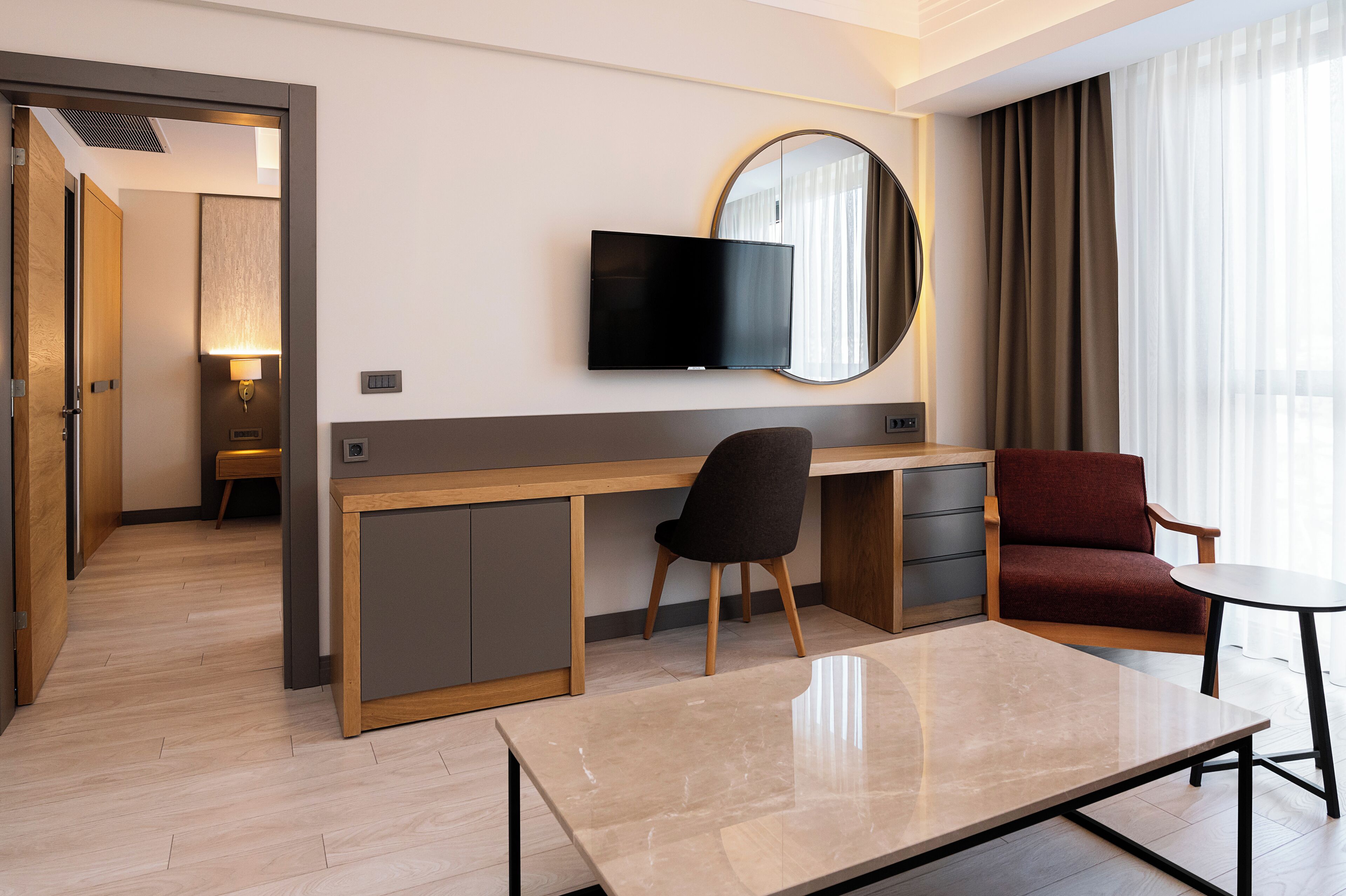 A hotel room with a wooden desk, a television mounted on the wall, a round mirror, a red sofa, and a small round table.