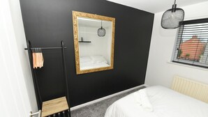 Un dormitorio con una pared negra, un espejo con marco dorado, una cama con ropa blanca, un radiador y una ventana con persianas.