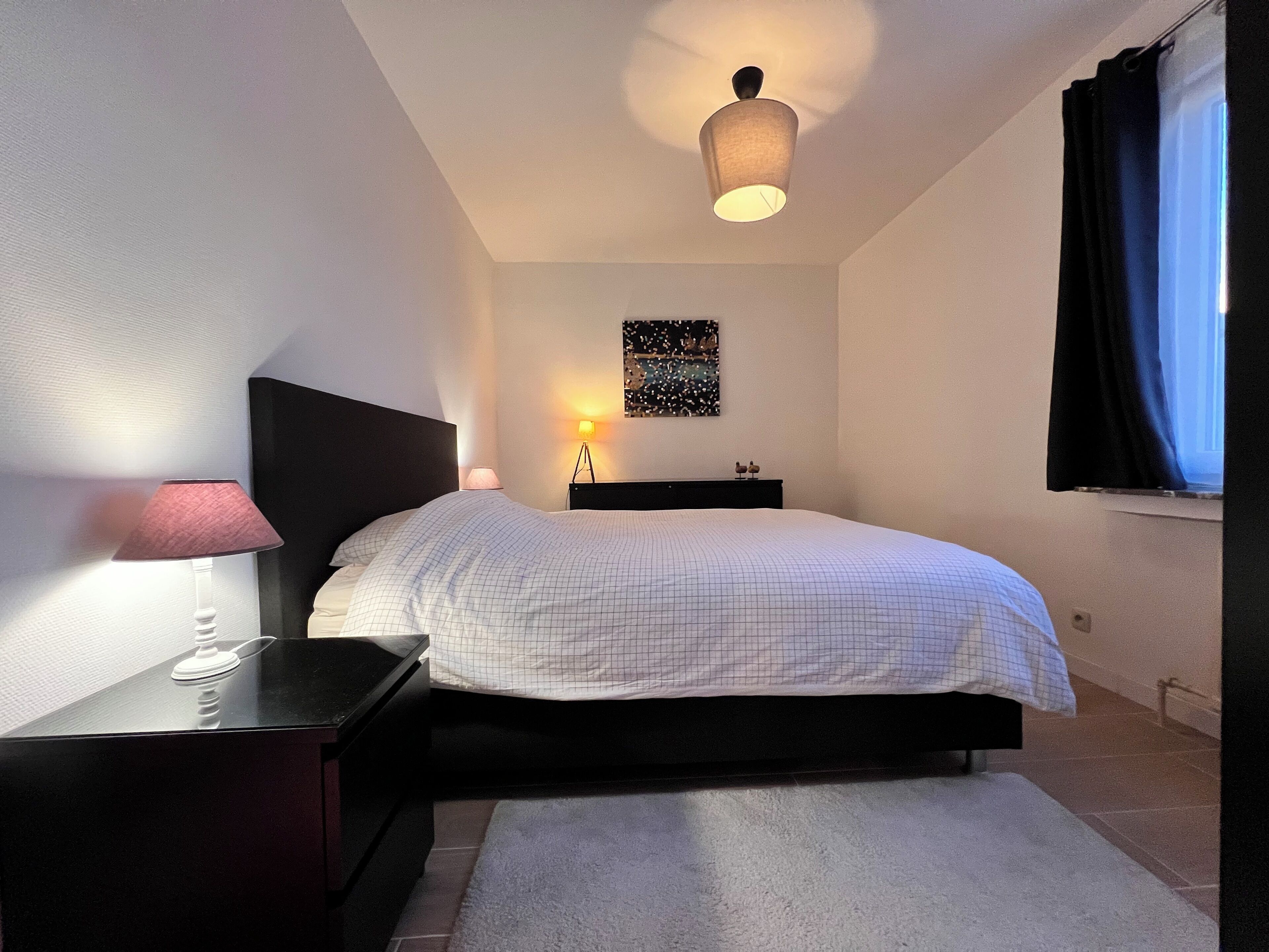 Ein Schlafzimmer mit Bett, Nachttischen, einer Lampe, Deckenleuchte, einem Fenster mit Vorhängen und einem Wandbild.