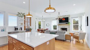 A modern kitchen with a large island, wooden cabinets, and pendant lighting. There is a dining area with a table and chairs, and a living room with a sofa and a fireplace. - Hilltop Views, Dog Friendly, Deck, Fire Pit & Near Pedernales Falls (Johnson City)
