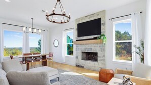 A spacious living room with a fireplace, a large TV, and a dining area with a view of the outdoors. - Hilltop Views, Dog Friendly, Deck, Fire Pit & Near Pedernales Falls (Johnson City)