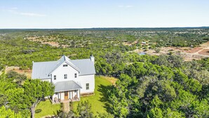 A large white house with a grey roof, surrounded by lush greenery and a clear blue sky. - Hilltop Views, Dog Friendly, Deck, Fire Pit & Near Pedernales Falls (Johnson City)