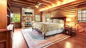 A log cabin bedroom with a stone fireplace, wooden ceiling beams, a large bed, and a nightstand with a lamp. - Mill Race Lodge I Waterfront, Pool, Wimberley (Wimberley)