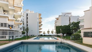 Una piscina rodeada de edificios modernos con balcones, cielo despejado y palmeras.