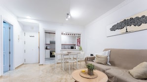 Una sala de estar con un sofá color beige, una mesa de centro con una planta, y un comedor con una mesa y sillas blancas.