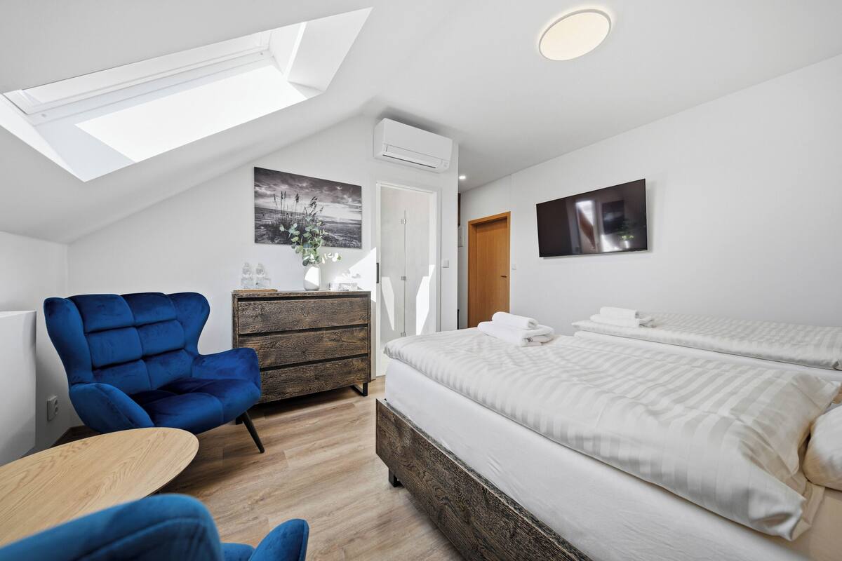 Una habitación de hotel moderna con tragaluz, una cama grande, un sillón azul, un aparador de madera y un televisor de pantalla plana.