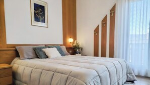 Een slaapkamer met een groot bed, houten lambrisering, een nachtkastje, een wandlamp en een raam met dunne gordijnen.