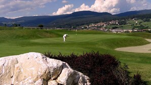 Een golfbaan met een grote rots op de voorgrond, een golfer in beweging en een stad verderop.
