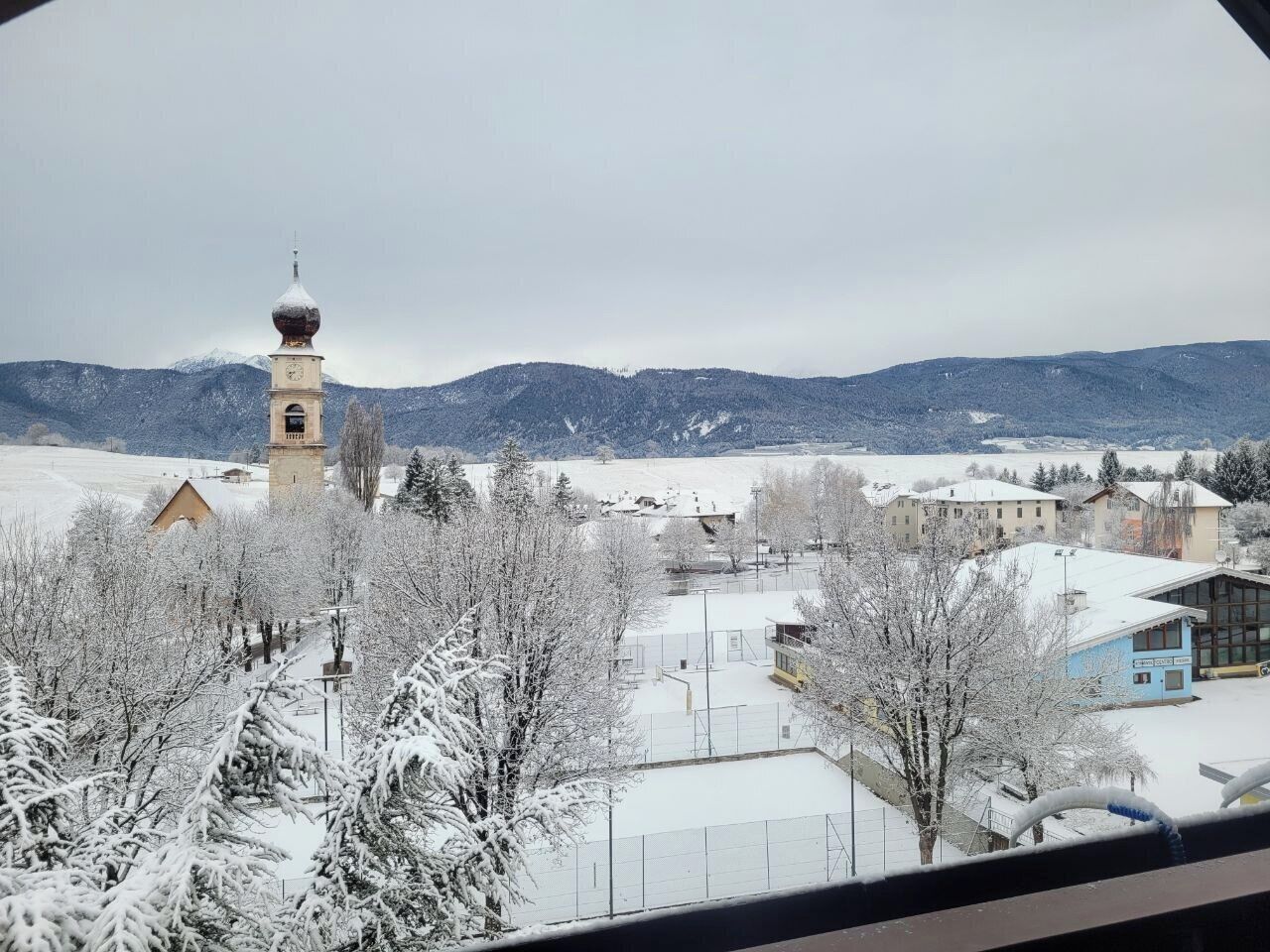 A snowy landscape with a church tower, buildings, and mountains in the background.