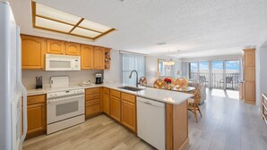 A modern kitchen with wooden cabinets, a microwave, stove, and dishwasher. There is a dining area with a table and chairs, and a balcony with a view.