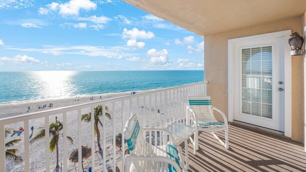 A balcony with a view of the beach and ocean, white chairs, a door with a window, and a palm tree.