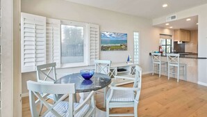Un coin repas avec une table ronde, des chaises blanches et un saladier bleu. On y trouve également un coin cuisine avec des placards en bois et une fenêtre équipée de stores.