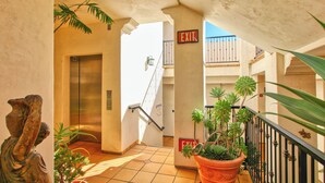 Un pasillo de hotel con ascensor, señal de salida, plantas en macetas y una escalera.