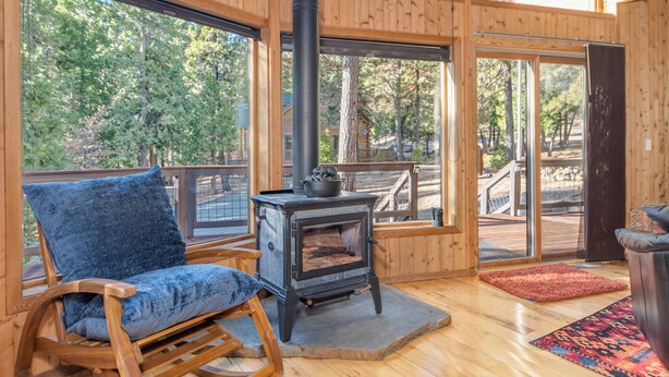 Interior acogedor de una cabaña con un sillón de madera mecedora, un estufa de leña y ventanas amplias que dan vista al bosque circundante.