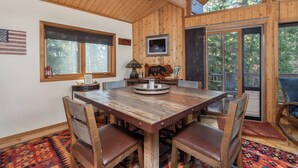 Una mesa de madera para comer con sillas, un ventilador de techo y un ventanal con vista a árboles.