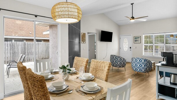 Un comedor con una mesa de madera dispuesta para cuatro personas, rodeada de sillas de mimbre, un ventilador de techo y una lámpara colgante grande.
