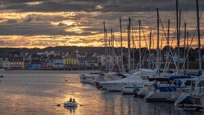 Un port de plaisance au coucher du soleil, avec des bateaux amarrés et une rangée de bâtiments colorés en arrière-plan.