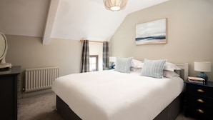 A bedroom with a large bed, a bedside table, a lamp, a radiator, and a window with curtains. - Two bedroom Cottage in the heart of Beadnell (Beadnell)