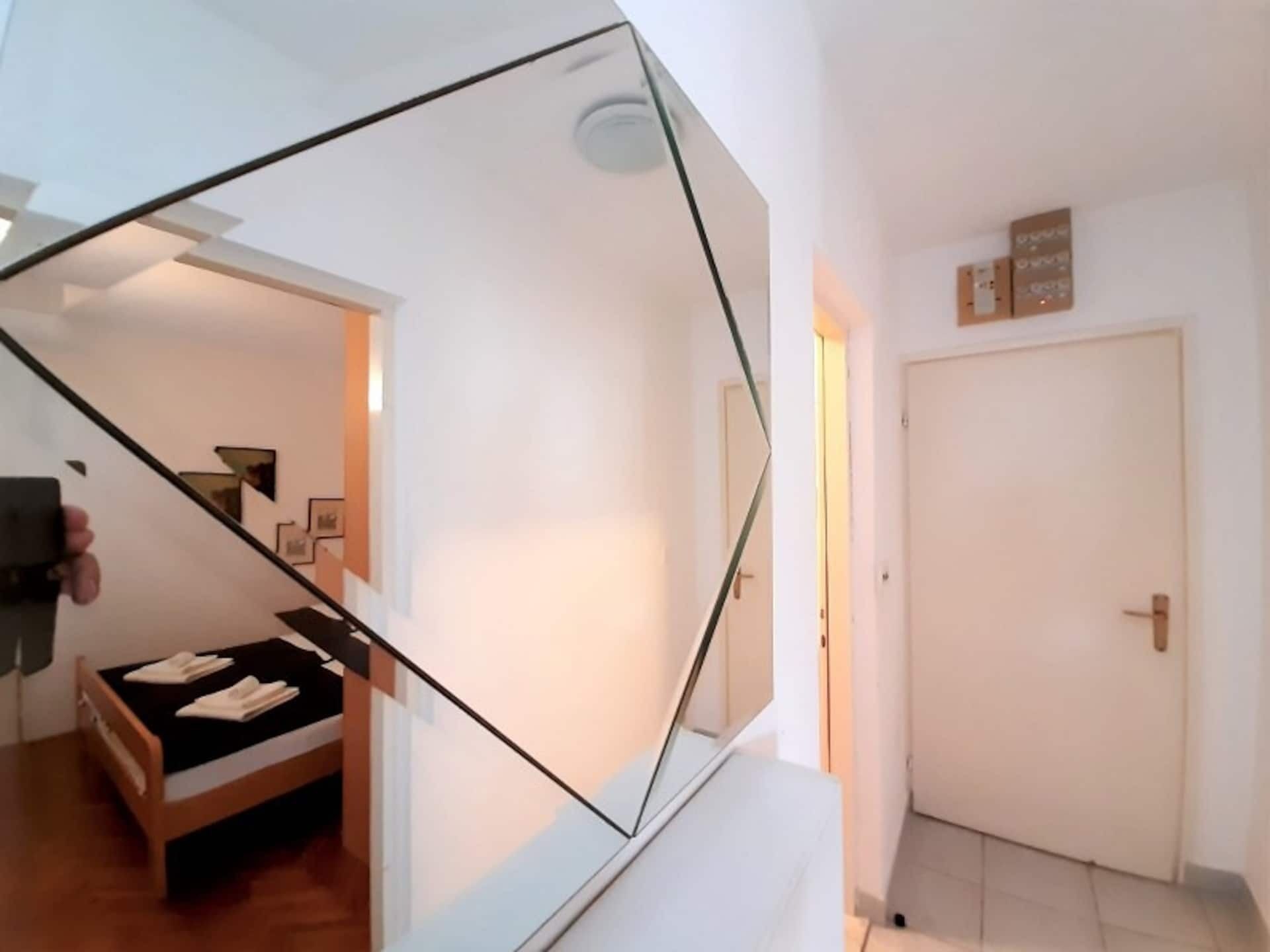 Ein modernes Interieur mit einer Spiegelwand, die ein Schlafzimmer, eine weiße Tür und ein kleines Wandregal reflektiert.