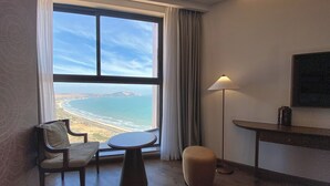 A hotel room with a large window offering a view of the beach and ocean. There is a chair, a small table, and a lamp.