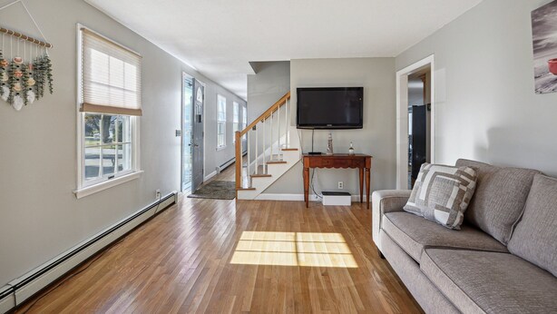 A spacious living room with hardwood flooring, a grey sofa, a wooden coffee table, and a flat-screen TV mounted on the wall. - 5 Bedroom Beach Retreat (Narragansett)