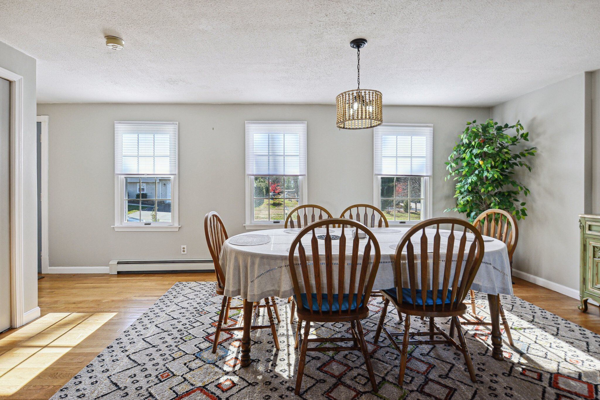 Une salle à manger avec une table ronde, six chaises en bois, un tapis à motifs et trois fenêtres équipées de stores.