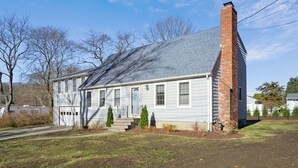 A single-story house with a brick chimney, a front garage, and a small front yard with a few trees and shrubs. - 5 Bedroom Beach Retreat (Narragansett)