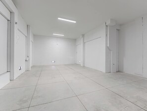 A spacious, empty room with white walls and a tiled floor, featuring two doors and a ceiling-mounted light fixture.